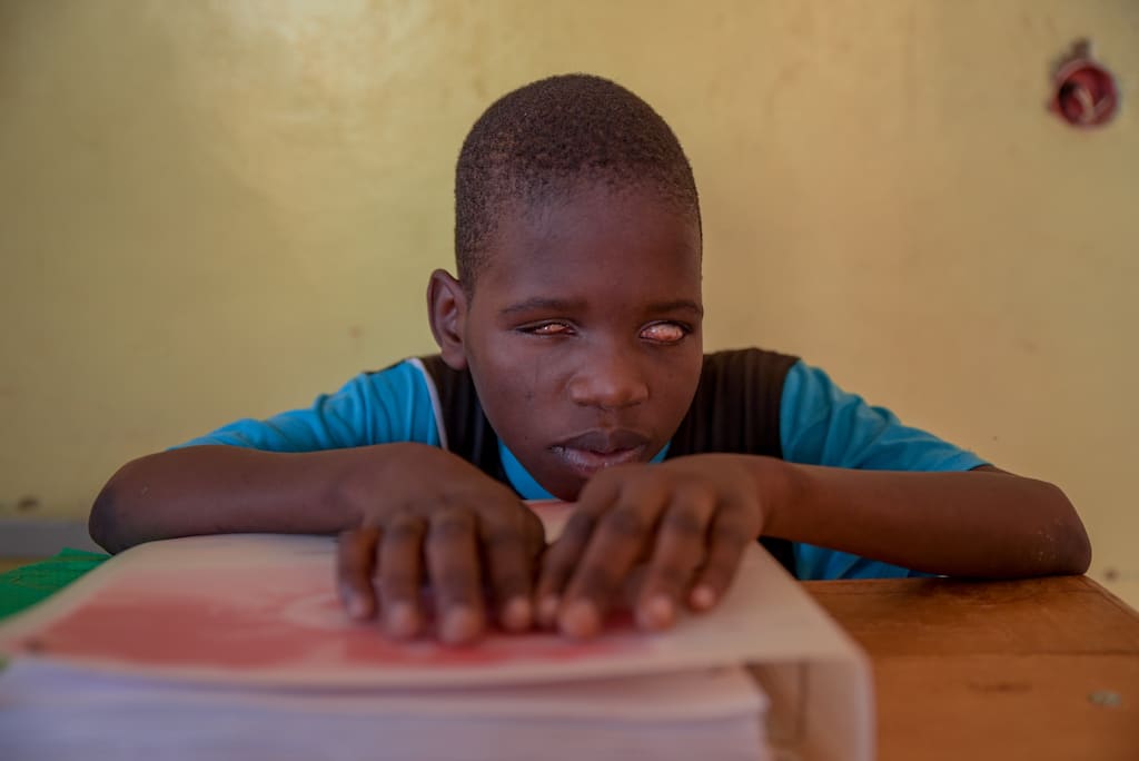 Kader is wearing a blue and black shirt. He is sitting at a desk in his classroom and has his hands on a Braille Bible in front of him. The wall behind him is yellow.